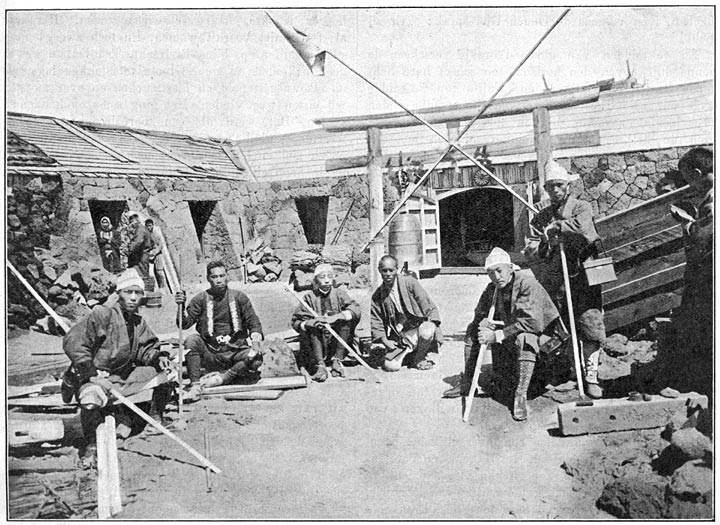 Binnenplaats van den tempel op den top van den Fuji. Op den achtergrond de heilige poort, waar men den stempel der priesters ontvangt.
De 2de man links op de afbeelding is de koelie “de ware duivel”, die op &eacute;&eacute;n jaar 45 keer den Fuji besteeg.